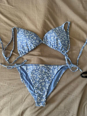 Blå blommig bikini från Kappahl  - Supersöt blå bikini från Kappahl i storlek XS med små vita blommor. Både överdel och underdel har knytband i sidorna för perfekt passform. Perfekt för lata dagar på stranden eller poolhäng med vänner. Toppen är i stl M. 
