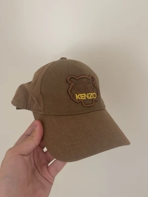 Brun Kenzo keps med tigerlogga - Snygg brun keps från Kenzo med broderad tigerlogga framtill och Kenzo-text baktill i gult. Kepsen har böjd skärm och justerbart spänne bak. Tillverkad i slitstarkt tyg, perfekt för en cool och avslappnad stil.