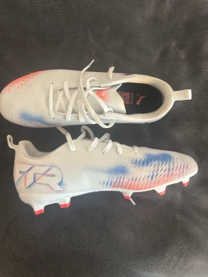 Vita Puma fotbollsskor med blå och röda detaljer -  Nya utan prislapp!!Säljer ett par snygga vita Puma fotbollsskor med blå och röda detaljer samt snörning. Skorna har en modern design med mönstrade inslag på sidorna och dobbar i rött. Perfekta för dig som vill sticka ut på planen!