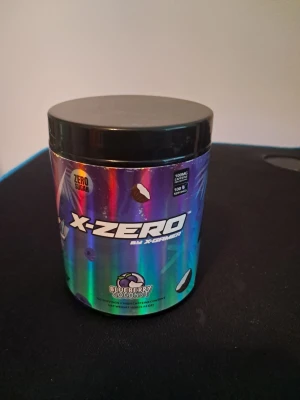 X-Zero Blueberry Coconut energidryck - Säljer en burk X-Zero energidryckspulver med smak av blåbär och kokos. Burken har en holografisk design med svart lock och innehåller 100 mg koffein per portion. Sockerfri och enkel att blanda med vatten för en uppiggande dryck.Produkten innehåller 100 portioner. 