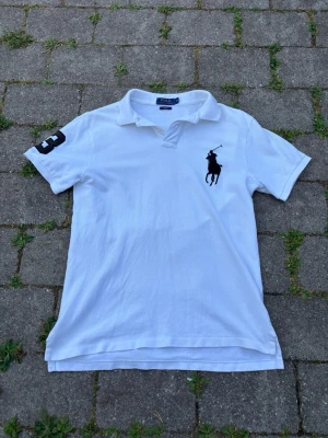 Vit pikétröja från Polo Ralph Lauren - Vit pikétröja från Polo Ralph Lauren i slim fit-modell. Tröjan har klassisk krage, korta ärmar och stor svart logga på bröstet samt siffra på ärmen. Perfekt för en clean och sportig stil.
