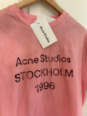 Rosa Acne Studios t-shirt med tryck - Säljer en rosa t-shirt från Acne Studios med svart texttryck 'Acne Studios STOCKHOLM 1996' på bröstet. Klassisk rund hals och korta ärmar. Perfekt för dig som gillar streetstyle och vill sticka ut med en ikonisk look.