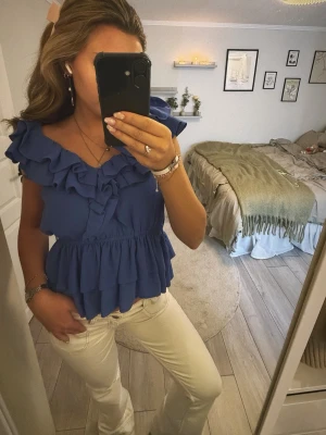 Blå volangblus med v-ringning - Supersöt blå blus med volanger och v-ringning. Toppen har ärmlös design och flera lager volanger både vid axlar och nederkant. Perfekt för dig som vill ha en feminin och trendig look. Tillverkad i ett lätt och luftigt material.