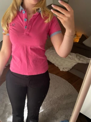 Rosa Fred Perry piké med blå detaljer - Säljer en snygg rosa pikétröja från Fred Perry med ljusblå krage och knappar. Tröjan har korta ärmar med matchande blå kant och den klassiska Fred Perry-loggan broderad på bröstet. Helt oandvänd och nypris 1000kr. Är en storlek M men passar de som har en storlek S 