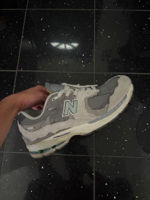 Grå New Balance 2002R sneakers - Säljer ett par grå New Balance 2002R sneakers med vit sula och vita skosnören. Skorna har en blandning av mocka och meshmaterial, klassisk N-logga på sidan och retroinspirerad design. Perfekta för dig som gillar stilrena och bekväma sneakers.