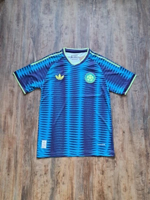 Blå sporttröja med mönster - Colombia  - Säljer en blå sporttröja med zigzag-mönster i mörkblått och ljusblått. Tröjan har korta ärmar, limegröna detaljer och tryck på bröstet. Tillverkad i ett lätt och ventilerande material, perfekt för träning eller match. Storlek: Står (M) men pasar som (S)