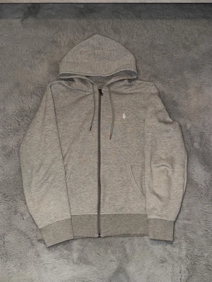  Polo Ralph Lauren zip hoddie  - Snygg grå hoodie med dragkedja från Polo Ralph Lauren. Klassisk design med huva och dragsnören, två fickor framtill och ribbade muddar. Liten vit logga broderad på bröstet. Perfekt för en chill och stilren look.