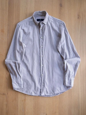Randig skjorta från Hansen & Jacob, size M - Säljer en klassisk långärmad skjorta från Hansen & Jacob med smala vertikala ränder i vitt och beige. Skjortan har knappar hela vägen, krage och är tillverkad i bomull. Perfekt för en clean och stilren look.