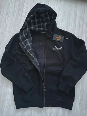 Svart hoodie med rutigt foder - Svart hoodie från Leon Marcel med dragkedja och rutigt foder i huvan. Broderad logga på bröstet och stort emblem med lejon och krona på ryggen. Mjuk och skön, perfekt för chill dagar. Snygga detaljer och klassisk passform.