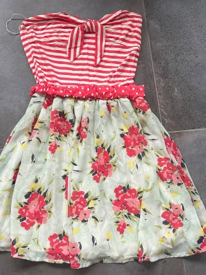 Randig och blommig sommarklänning - Säljer en söt axelbandslös klänning med rödvita ränder upptill och en rosett framtill. Kjolen är vit med blommigt mönster i rött, rosa och gult. Midjan har en prickig detalj. Perfekt för varma dagar och ger en riktigt somrig känsla.