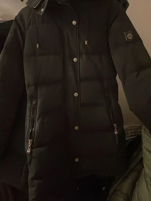 Svart dunjacka med huva och dragkedjor - Snygg svart dunjacka från Save The Duck med huva och silverfärgade dragkedjor på fickorna. Jackan har quiltad design, långa ärmar och tryckknappar framtill. Perfekt för kalla dagar och enkel att matcha med allt. Den har används några gånger men går att tvätta! Pris kan diskuteras.