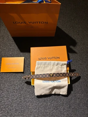 Louis Vuitton  armband - Snyggt armband från Louis Vuitton i klassiskt brunt monogrammönster med guldfärgad V-detalj framtill. Tillverkat i canvas och läder, perfekt för dig som gillar exklusiva accessoarer. Kommer med originalpåse, ask och påse från Louis Vuitton.