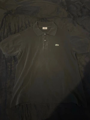 Svart pikétröja från Lacoste - Klassisk svart pikétröja från Lacoste med den ikoniska gröna krokodilloggan på bröstet. Tröjan har korta ärmar, krage och knappar framtill. Tillverkad i bomull för en skön och luftig känsla.