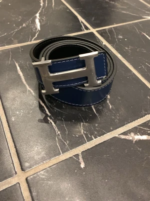 Blått skärp med stor H-spänne - Snyggt blått skärp med en stor, silverfärgad H-formad metallspänne. Skärpet har vita sömmar längs kanterna och är gjort i slätt material som ger en stilren look. Perfekt accessoar för att lyfta din outfit.