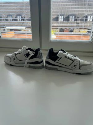 Vita och svarta Louis Vuitton sneakers - Säljer ett par stilrena Louis Vuitton sneakers i vitt och svart med klassisk LV-logga på sidan och tungan. Skorna har en chunky sula och detaljer i både läder och textil. Perfekta för dig som vill ha en exklusiv och trendig look.