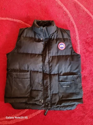 Svart dunväst från Canada Goose - Snygg svart dunväst från Canada Goose med hög krage och två stora fickor framtill. Västen har ett klassiskt Canada Goose-märke på bröstet och stängs med både dragkedja och knappar. Perfekt för lager-på-lager och håller dig varm under kyliga dagar.