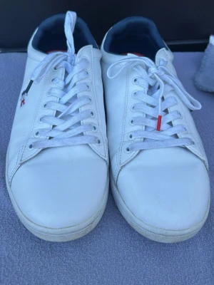 Vita lsneakers Lacoste  - Snygga vita sneakers ifrån Lacoste  läder med klassisk låg profil och vita skosnören. Skorna har små röda och svarta detaljer på snörningen och en mörkblå insida. Perfekta för en clean och stilren look.  Stl 43