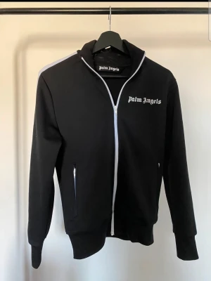 Svart hoodie från Palm Angels - Snygg svart hoodie från Palm Angels med vit dragkedja och logga på bröstet. Hoodien har långa ärmar, två fickor med dragkedja och en klassisk passform. Materialet är mjukt och bekvämt, perfekt för en streetwear-look.