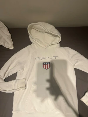 Vit hoodie från GANT med logga - Säljer en vit hoodie från GANT med broderad logga och sköld på bröstet. Hoodien har en klassisk huva och en stor magficka framtill. Tillverkad i mjukt material och har ribbade muddar vid ärmar och nederkant. Perfekt för en avslappnad och stilren look.
