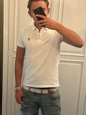 Pikétröja Lyle & Scott - Vit pikétröja från Lyle & Scott med klassisk krage, knappar framtill och den ikoniska gula loggan på bröstet. Tillverkad i bomull för en skön och luftig känsla. Enkel och stilren modell som funkar till det mesta. Storlek:14-15/xs. Lite sliten på insidan av kragen.
