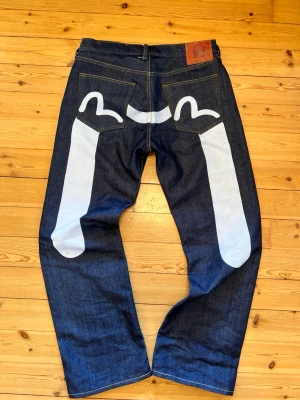 Raw denim selvage baggy evisu jeans - Unika mörkblå jeans med bred passform och stor vit grafisk målning på baksidan. Jeansen har klassiska bakfickor och en läderpatch i midjan med texten 'No 2'. Materialet är kraftig denim i 100% bomull.