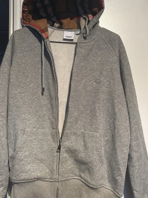 Grå zip hoodie från Burberry - Säljer en grå zip hoodie från Burberry med klassiskt rutigt foder i huvan och dragsko. Hoodien har fickor framtill och diskret Burberry-logga broderad på bröstet. Perfekt för dig som gillar stilrena och exklusiva plagg. Däremot gick dragkedjan sönder så den är lagad med en ny rejäl