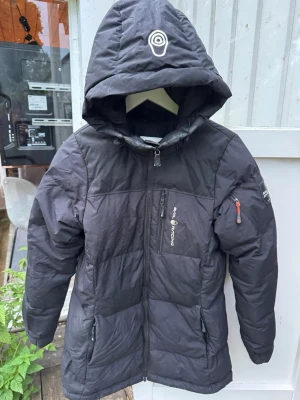 Svart pufferjacka från Sail Racing - Svart pufferjacka från Sail Racing med huva och dragkedja framtill. Jackan har två sidofickor med dragkedja, en bröstficka och en ärmficka med orange dragkedja. Perfekt för kalla vinterdagar och har en sportig look.