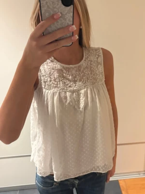 Vit ärmlös blus med spetsdetaljer - Säljer en luftig vit blus med broderade spetsdetaljer upptill och prickigt mönster i tyget. Blusen är ärmlös och har en lös passform som ger en avslappnad och somrig känsla. Perfekt att matcha med jeans eller kjol för en fräsch look.