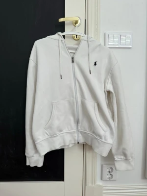 Vit zip hoodie med logga - Snygg vit hoodie med dragkedja och huva. Klassisk design med två fickor framtill och justerbara snören i huvan. Liten svart logga broderad på bröstet. Tillverkad i mjukt material som känns skönt mot huden. Perfekt för en avslappnad och stilren look.