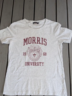 Grå Morris University t-shirt - Snygg ljusgrå t-shirt från Morris med tryck i vinrött framtill där det står 'Morris University 1968'. Klassisk rund halsringning och korta ärmar. Perfekt för en avslappnad och stilren look. Trycket är något nött.