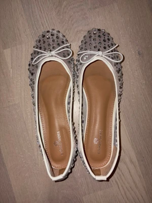Vita mesh ballerinaskor med strass - Säljer ett par vita ballerinaskor i mesh med glittrande strass och små rosetter framtill. Skorna har vita kanter och en mjuk innersula i beige. Perfekt för dig som gillar detaljer och vill ha något extra till din outfit.