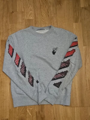 Grå hoodie med grafiska röda detaljer - Snygg grå hoodie med unika grafiska röda och svarta detaljer på ärmarna och stor printad design på ryggen. Crewneck-modell med ribbade muddar och Off-tryck på bröstet. Perfekt för dig som gillar streetwear och vill sticka ut.