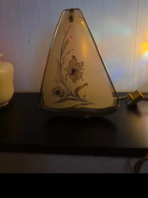 Triangelformad retro vägglampa i glas - Retro vägglampa i triangelformad design med genomskinligt glas och blommönster i svart. Lampan har en guldfärgad kant och strömbrytare på sladden. Perfekt som dekorativ belysning i rummet.