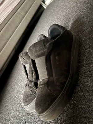 Svarta sneakers från Axel Arigato - Stilrena svarta sneakers från Axel Arigato i mocka med diskret logga på sidan och gummisula. Klassisk low-top modell utan snörning, med elastiska band över vristen för enkel påtagning. Perfekta för en clean och modern look.