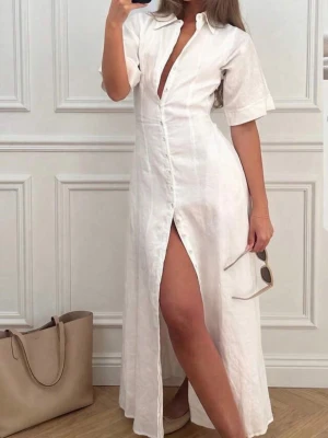 Vit långklänning i linne med slits - Stilren vit långklänning  i linne med knappar framtill, korta ärmar och en djup v-ringning. Klänningen har en hög slits som ger en trendig look och passar perfekt till sommaren. Enkel och elegant design som är lätt att styla.