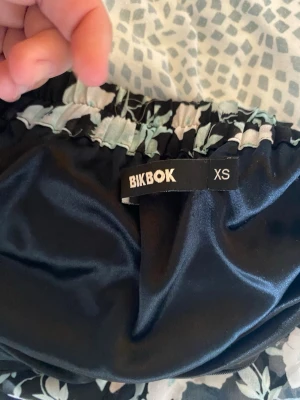 Blommig svart kjol från BikBok - Säljer en svart kjol från BikBok i storlek XS med ljusblått blommönster. Kjolen har resår i midjan och är gjord i ett mjukt, glansigt material som känns skönt mot huden. Perfekt för dig som gillar en feminin och trendig stil.