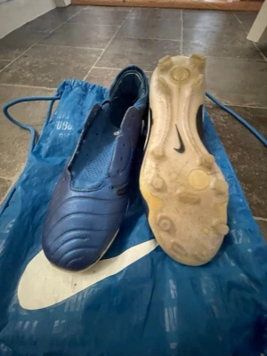 Blå Nike fotbollsskor med dobbar - Säljer ett par blå Nike fotbollsskor med klassisk design och dobbar för bra grepp på planen. Skorna har en slät ovandel i syntetmaterial och en vit sula med svarta detaljer. Perfekta för dig som vill ha stilrena och funktionella skor till match eller träning.