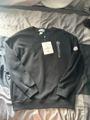 Moncler tröja  - Svart sweatshirt från Moncler med rund halsringning och vit broderad Moncler-logga på bröstet. Klassisk passform med ribbade muddar vid ärmslut och nederkant. Moncler-märke på vänster ärm. Perfekt för en clean och stilren look.