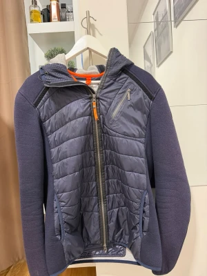 Blå hybridjacka från Parajumpers - Snygg blå hybridjacka från Parajumpers med quiltad front och huva. Jackan har dragkedja, bröstficka med zip och kontrasterande material på ärmar och sidor. Perfekt för lager-på-lager och streetwear-vibe.