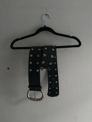 Svart bälte med silvernitar - Snyggt svart bälte med dubbla rader silverfärgade nitar och stor silverspänne. Perfekt accessoar för att ge din outfit en edgy touch. Bältet är i syntetmaterial och passar till många olika stilar.