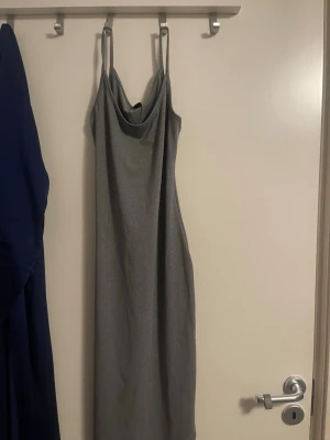 Grå långklänning med smala band - Säljer en elegant grå långklänning med smala axelband och draperad urringning. Klänningen har en figurnära passform och skimrande material som ger en lyxig känsla. Perfekt för dig som vill sticka ut med en stilren look.
