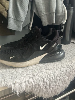 Nike Air Max 270 svarta sneakers - Svarta Nike Air Max 270 sneakers med vit sula och tydlig Air-enhet i hälen. Skorna har mesh-överdel för bra ventilation, vita detaljer och logga på sidan. Snygg och sportig design som passar till streetwear och casual outfits.