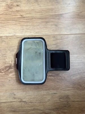 Svart sportarmband för mobil - Praktiskt svart sportarmband för mobiltelefon med transparent plastfönster och justerbart kardborreband. Perfekt för träning och löpning, tillverkat i mjukt och flexibelt material som sitter bekvämt runt armen. Passar de flesta smartphones.