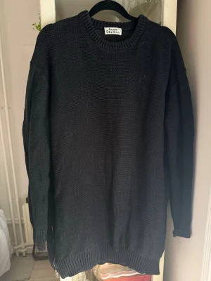 Svart stickad tröja från Acne Studios - En svart, oversized stickad tröja från Acne Studios med rund halsringning och ribbade muddar vid ärmslut och nederkant. Tröjan har en minimalistisk design och är perfekt för lager-på-lager-stil. Dragkedja på sidorna. 