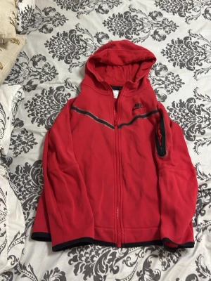 Röd Nike hoodie med dragkedja - Snygg röd hoodie från Nike med hel dragkedja framtill och svart detalj över bröstet. Huva och muddar i svart ger en sportig look. Perfekt för dig som gillar streetwear och vill ha något bekvämt och stilrent. 158 cm