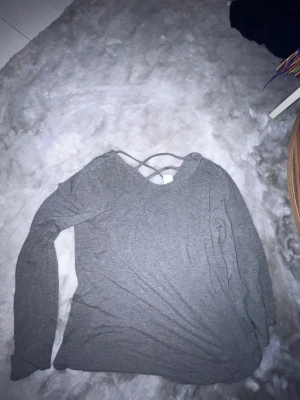 Grå långärmad topp med snörning - En enkel och stilren grå långärmad topp med snygg snörningsdetalj i ryggen. Perfekt att matcha med jeans eller kjol för en avslappnad look. Tillverkad i mjukt material som känns skönt mot huden.