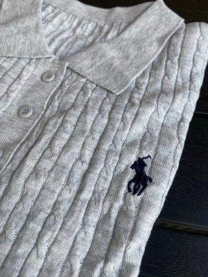 Vit kabelstickad pikétröja Polo Ralph Lauren - Vit kabelstickad pikétröja från Polo Ralph Lauren med korta ärmar, klassisk krage och knappar framtill. Tröjan har en broderad marinblå logga på bröstet och ribbade muddar vid ärmslut och nederkant. Perfekt till en clean och stilren look.