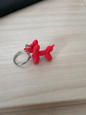 Nyckelring med röd ballonghund - Säljer en unik nyckelring med en röd ballonghund i plast. Nyckelringen har en metallring och ballonghunden är formad som en klassisk ballongfigur. Perfekt att hänga på väskan eller nycklarna för en lekfull touch.