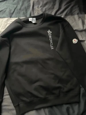  Moncler tröja - Snygg svart sweatshirt från Moncler med logga tryckt vertikalt på bröstet och ett Moncler-märke på ärmen. Tröjan har rund halsringning, ribbade muddar och är tillverkad i mjukt material som passar perfekt till kalla dagar.
