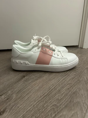 Valentino open - Fräscha vita sneakers med bred rosa rem över snörningen och coola nitar på sidan av sulan. Skorna har rund tå, vita skosnören och en chunky vit sula. Perfekta för dig som gillar en stilren men ändå trendig look.